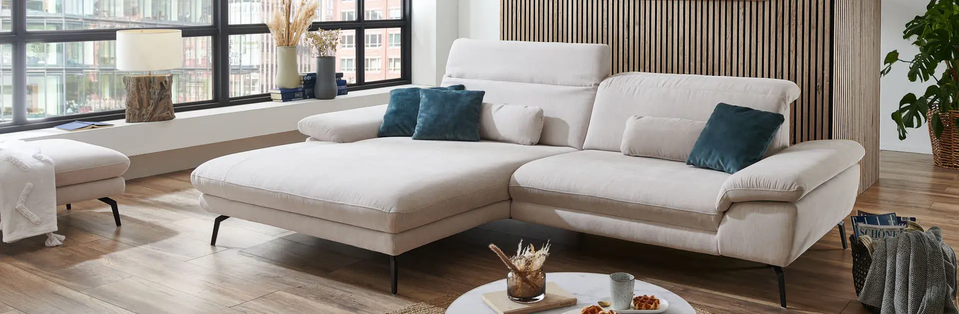 Modernes Wohnzimmer mit hellbeigem Ecksofa, blauen Zierkissen, Holzlamellenwand und großem Fenster mit Stadtblick